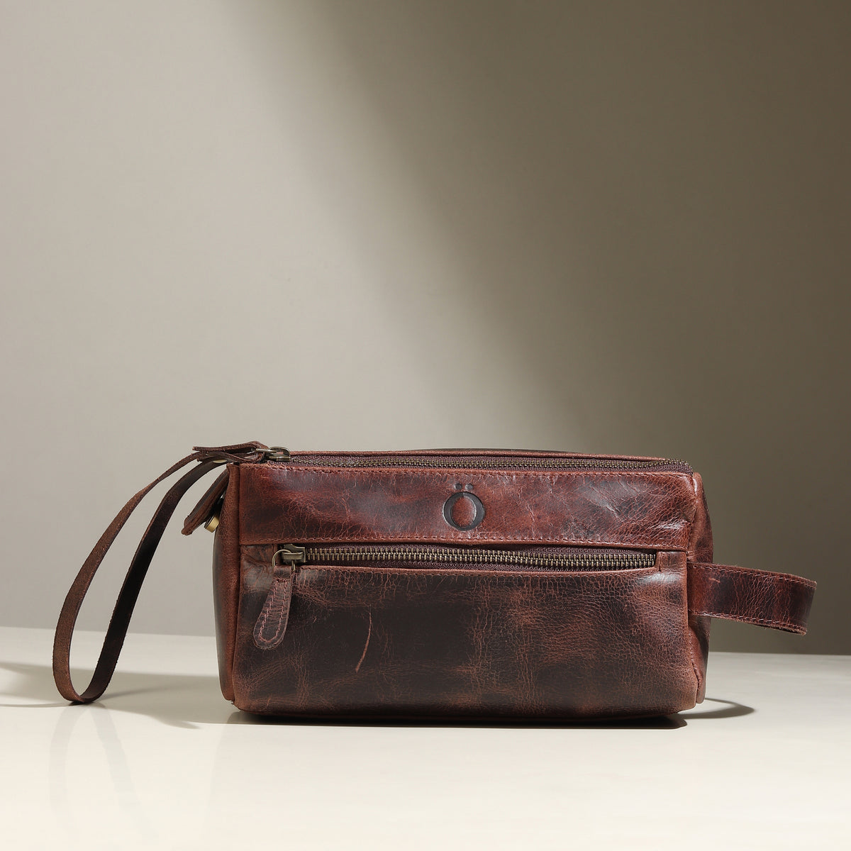 Front view of a closed dark brown leather wash bag with a zip top and wrist loop handle